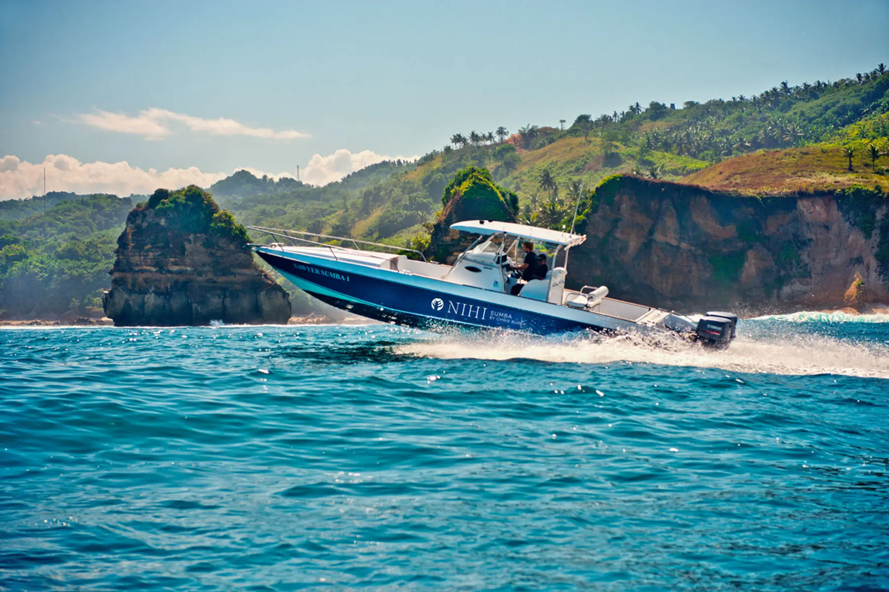 The 'Sumba Sawyer' boat navigating the clear waters off Sumba Island during a boat charter, with lush green hills and coastal cliffs in the background.