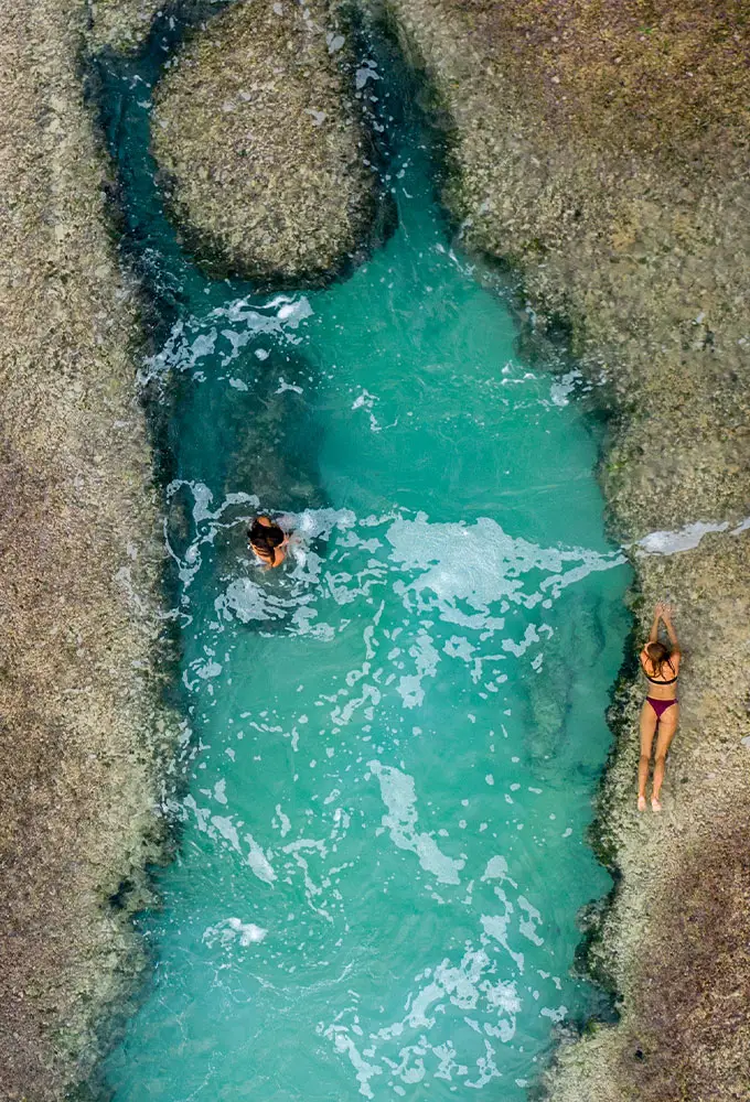 Two guests enjoying the clear turquoise water of a tidal rock pool at Cleopatra Rock during low tide, with one guest swimming and the other relaxing by the pool's edge at Nihiwatu Beach.