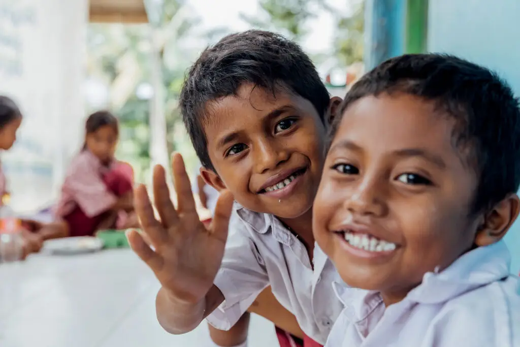 Two young Sumbanese boys in school uniforms smiling and waving during a classroom lunch session, reflecting the positive impact of educational programs supported by the Sumba Foundation and Burch Family Foundation for local communities.