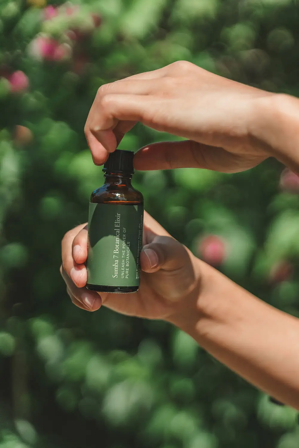 Hands opening a bottle of Sumba 7 Botanical Elixir, a natural skin treatment oil made with wild-harvested Indonesian botanicals, photographed against a lush green background at NIHI Sumba.