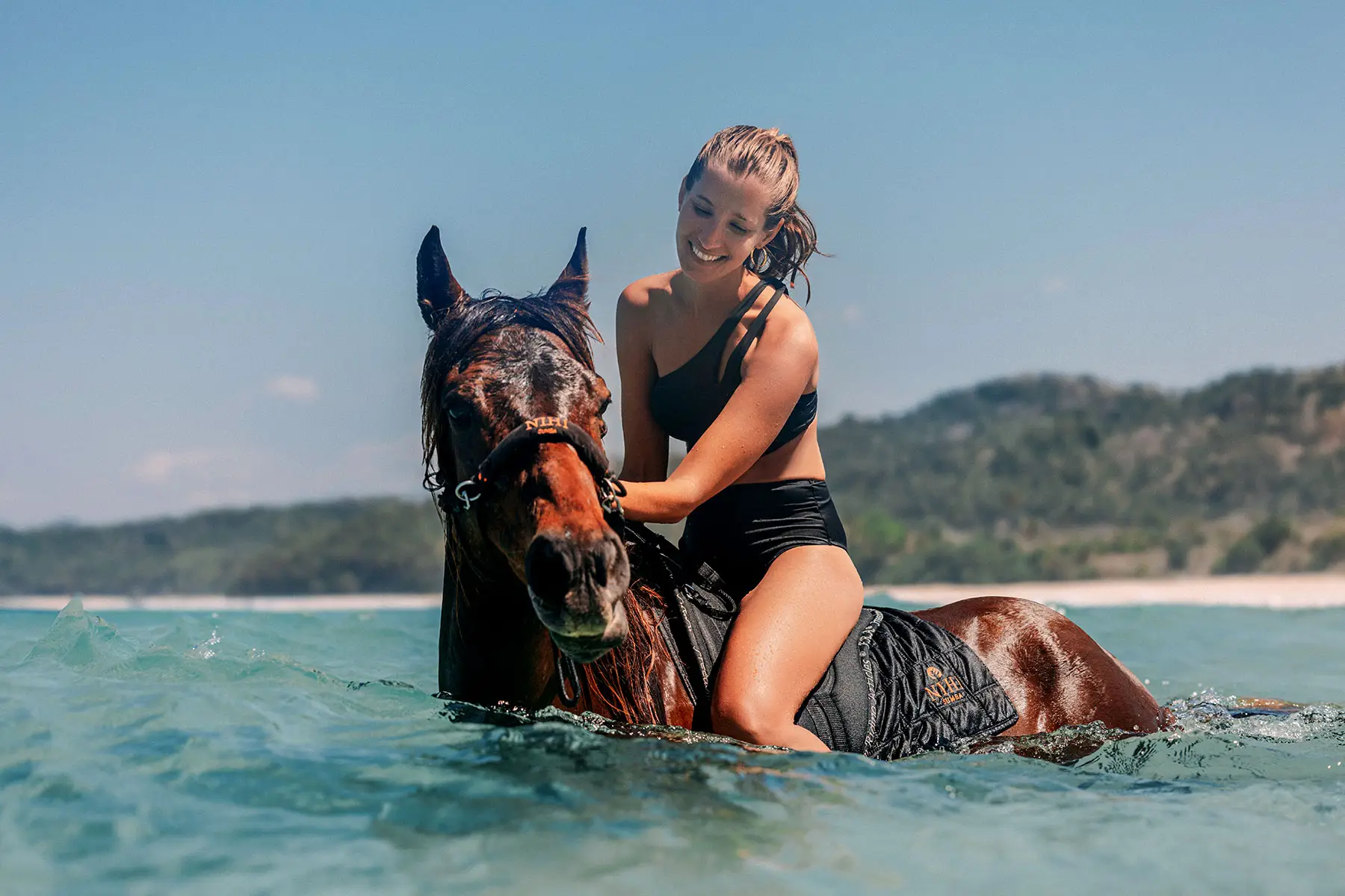 Smiling guest in swimwear riding a Sumbanese horse through the shallow ocean waters of Nihiwatu Beach at NIHI Sumba, guided by a local handler during the resort’s iconic swimming with horses experience.