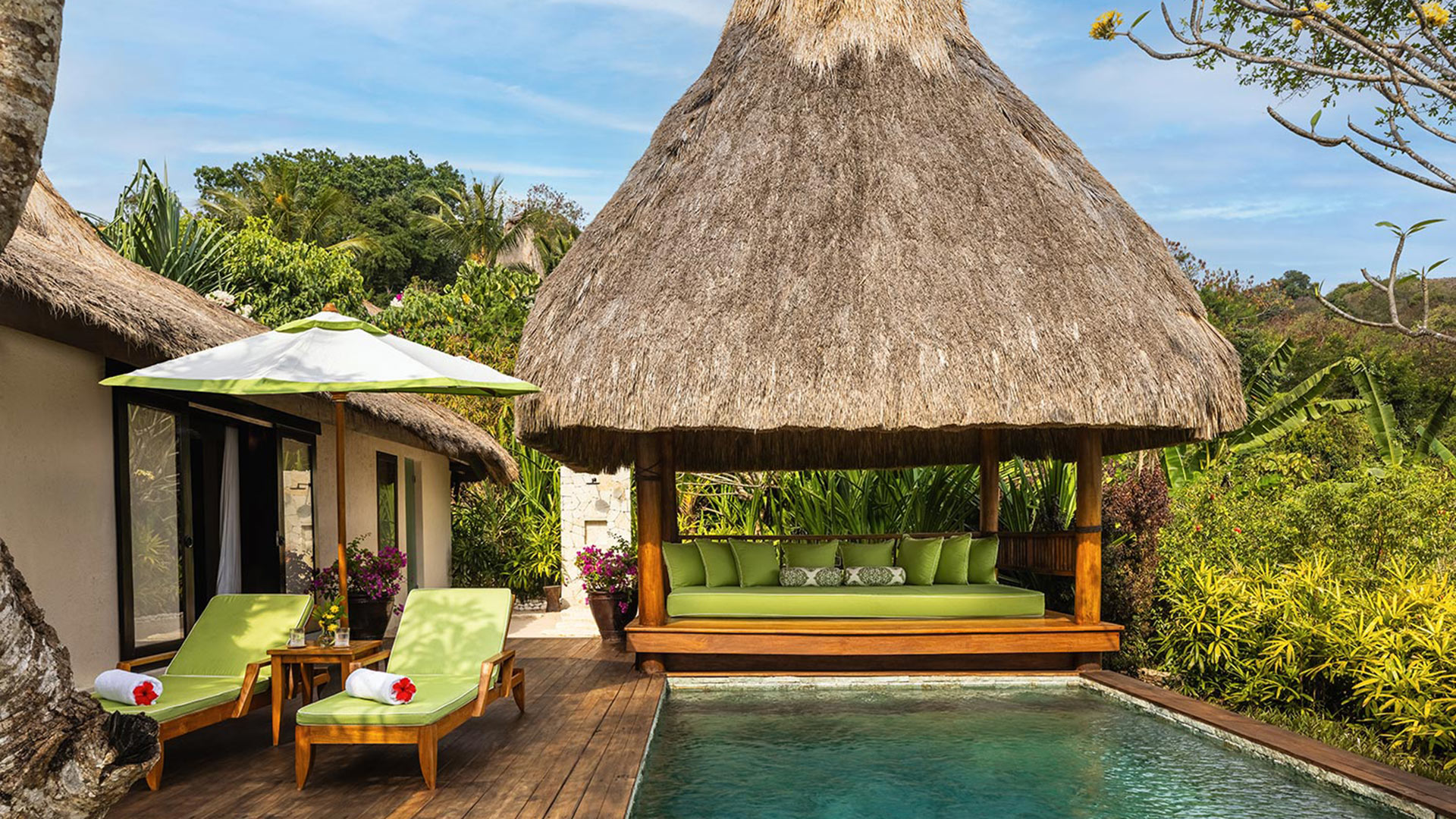 Tranquil outdoor lounge area at Lulu Amahu Saudara Villa, NIHI® Sumba, featuring a thatched gazebo with plush green seating, an umbrella, and a private plunge pool surrounded by lush tropical greenery and colorful flowers.