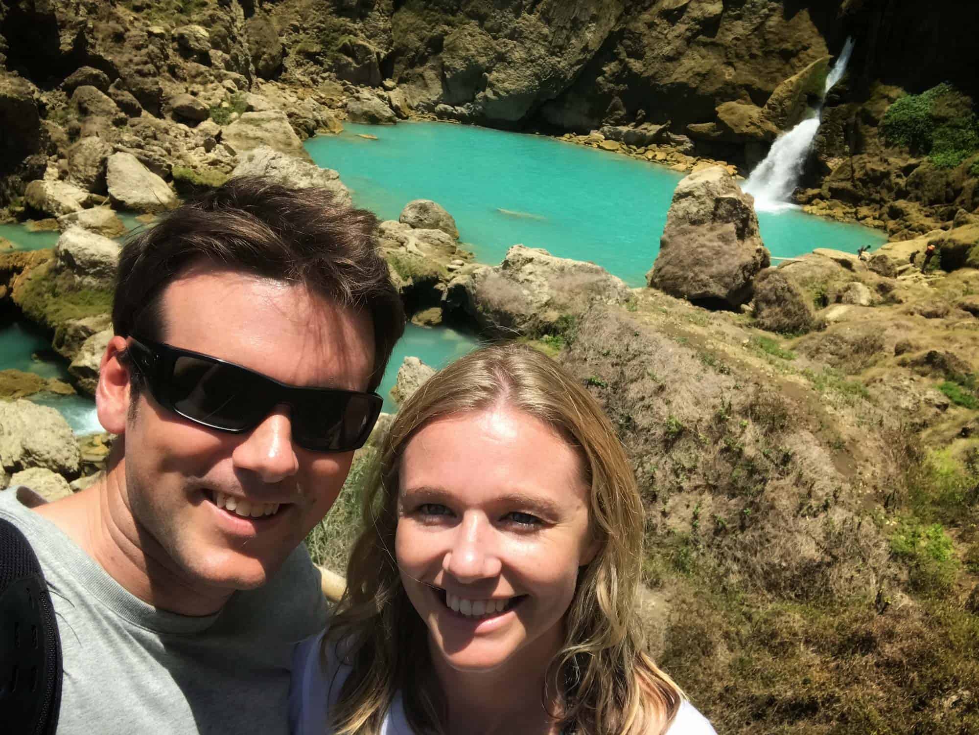 Happy couple taking a selfie at the stunning Blue Waterfall in Sumba, Indonesia, surrounded by dramatic cliffs and natural rock formations — highlighting romantic adventures and immersive nature experiences at NIHI Sumba.