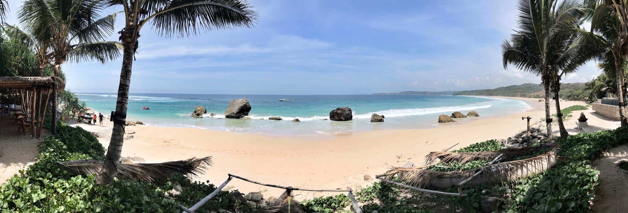 Panoramic view of the pristine beach at NIHI Sumba, Indonesia, showcasing crystal-clear turquoise waters, dramatic rock formations, and lush palm trees — a perfect setting for luxury travel and wellness retreats.