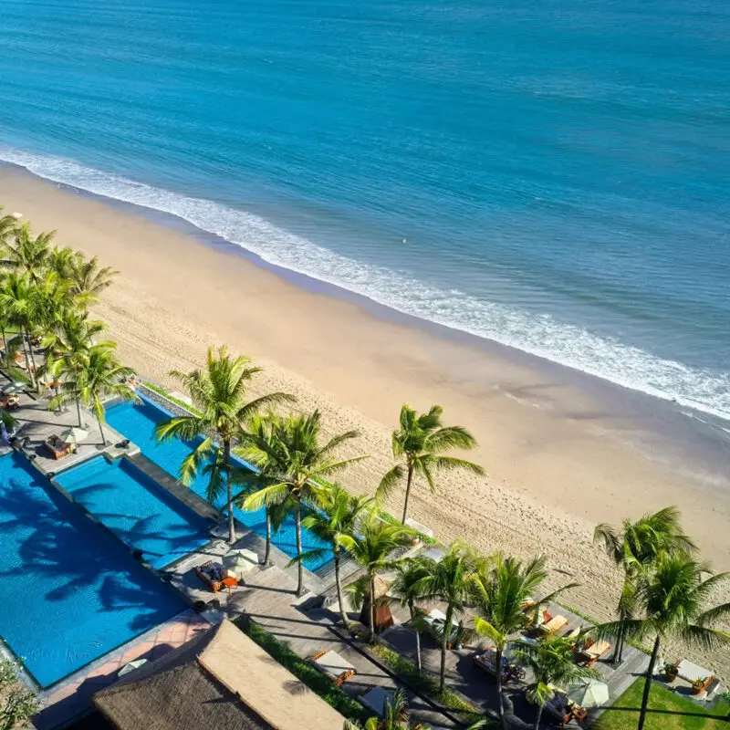 Aerial view of the pool at The Legian Seminyak, Bali—NIHI’s partner property—featuring a luxury beachfront setting with swaying palm trees, golden sands, and turquoise waters along Legian Beach.